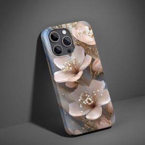 Puede incluir: Funda de teléfono con diseño floral. La funda tiene un fondo azul claro y gris con delicadas flores rosas y detalles dorados. Las flores tienen centros blancos con forma de perla, creando un efecto 3D. Diseñada para un smartphone.