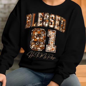 May include: Black sweatshirt with the word "BLESSED" in a plaid and floral design above the number "91". The phrase "He Is My Refuge" is written below the number. The sweatshirt is a casual style.