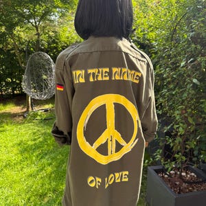 Könnte beinhalten: Olivgrüne Jacke mit dem Text "IN THE NAME OF LOVE" und einem gelben Peace-Zeichen auf dem Rücken. Die Jacke hat auch einen deutschen Flaggenaufnäher am Ärmel.