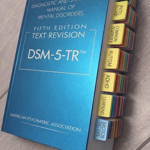 Könnte beinhalten: Ein blaues Buch mit dem Titel "Diagnostic and Statistical Manual of Mental Disorders, Fifth Edition, Text Revision, DSM-5-TR". Das Buch hat Registerkarten für verschiedene Störungen, darunter Autismus und ADHS. Herausgegeben von der American Psychiatric Association.