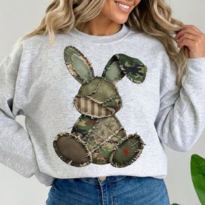 May include: A light grey sweatshirt with a patchwork bunny design. The bunny is made of various camouflage patterns and fabrics, with a star and cross detail. The sweatshirt is a casual, comfortable style.
