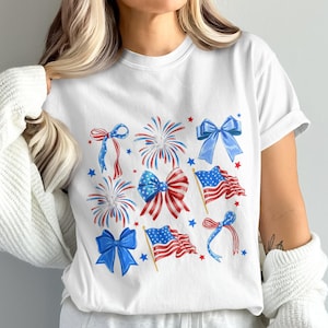 Puede incluir: Camiseta blanca con un diseño patriótico. El diseño incluye lazos, fuegos artificiales, banderas americanas y estrellas rojas, blancas y azules. El diseño es de estilo acuarela, ideal para eventos patrióticos.