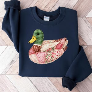 Könnte beinhalten: Marineblaues Sweatshirt mit einem Enten-Design im Patchwork-Stil. Die Ente hat einen grün karierten Kopf, einen gelben Schnabel und einen Körper aus verschiedenen Stoffmustern in Rosa, Rot und Braun. Das Sweatshirt hat einen Rundhalsausschnitt und lange Ärmel mit gerippten Bündchen.