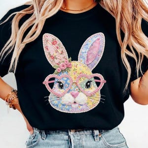 May include: Black t-shirt featuring a colorful bunny face with heart-shaped glasses. The bunny has floral patterned ears and a yellow, blue, and pink patchwork design. The glasses are pink, and the bunny has a pink nose.