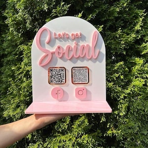 Könnte beinhalten: Ein rosa-weißes Schild mit dem Text "Let's get Social" in Schreibschrift. Das Schild hat zwei QR-Codes und Facebook- und Instagram-Symbole. Das Schild wird vor einem grünen, belaubten Hintergrund gehalten.
