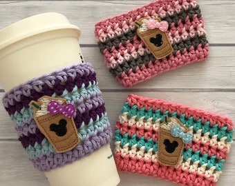 May include: Three crocheted coffee cup cozies with a Mickey Mouse design. The cozies are in pink, purple, and teal colors with white stripes. Each cozy has a brown felt applique of a Mickey Mouse head with a coffee cup and a pink bow.