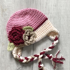 May include: A pink, beige, and burgundy crocheted baby hat with a braided cord and flower embellishments.