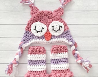 May include: A pink and purple crocheted owl hat and matching leg warmers. The hat has a white face with black eyes and a pink beak. The leg warmers have a pink and white striped pattern.