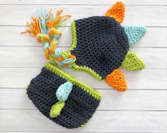 May include: A crocheted dinosaur hat and matching nappy cover set in navy blue with green, orange and light blue accents.