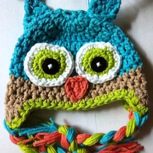 May include: A crocheted owl hat with a teal blue body, green and white eyes, and a brown, green, and orange brim. The hat has a long, braided tassel in teal blue, green, and orange.