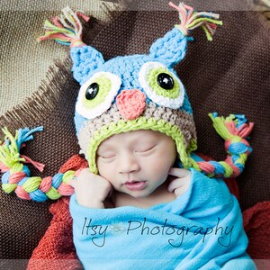 May include: A baby wearing a blue, green, and pink crocheted owl hat with a long braided tail. The hat has large, colorful eyes and a pink beak. The baby is sleeping peacefully on a brown burlap background.