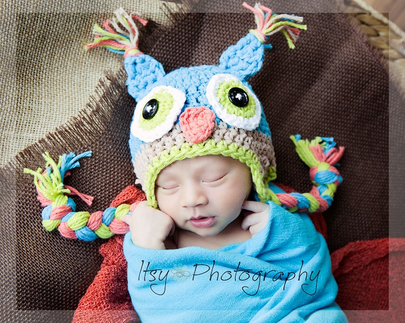 May include: A baby wearing a blue, green, and pink crocheted owl hat with a long braided tail. The hat has large, colorful eyes and a pink beak. The baby is sleeping peacefully on a brown burlap background.