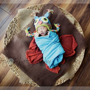 May include: A newborn baby wearing a blue and green owl hat with brown eyes and a yellow beak, wrapped in a blue blanket, lying in a woven basket with a brown burlap lining.