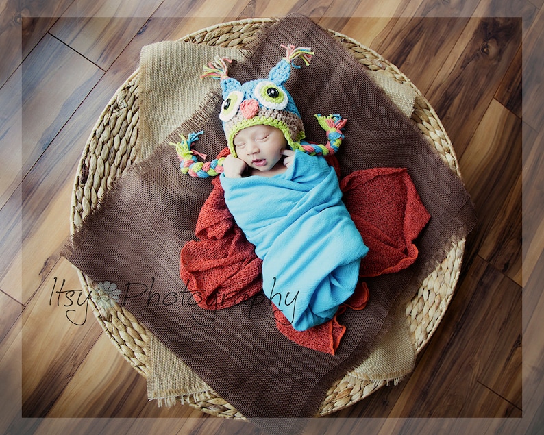 May include: A newborn baby wearing a blue and green owl hat with brown eyes and a yellow beak, wrapped in a blue blanket, lying in a woven basket with a brown burlap lining.