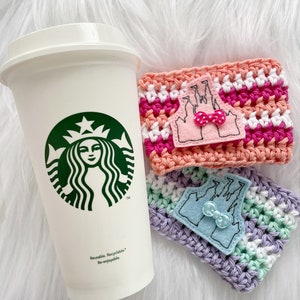 Pode incluir: Dois aquecedores de chávena de café de crochê com um design de castelo rosa e branco e um design de castelo azul e branco. Os aquecedores estão sobre uma superfície branca e fofa ao lado de uma chávena de Starbucks branca com o logotipo da Starbucks.