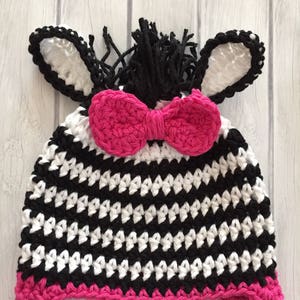 May include: A black and white striped crocheted zebra hat with pink ear flaps and a pink bow. The hat has a black mane and white ears.