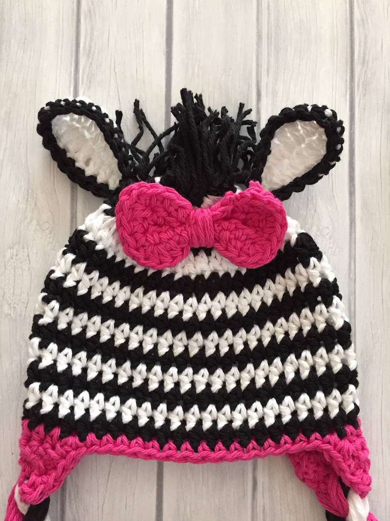 May include: A black and white striped crocheted zebra hat with pink ear flaps and a pink bow. The hat has a black mane and white ears.