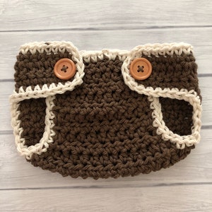 May include: A handmade, brown crochet diaper cover with a cream-colored trim and two wooden buttons. The diaper cover is designed with a textured stitch and is suitable for a baby. The buttons are used for closure.
