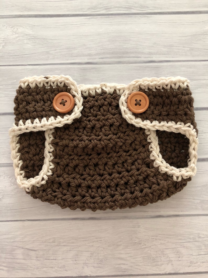 May include: A handmade, brown crochet diaper cover with a cream-colored trim and two wooden buttons. The diaper cover is designed with a textured stitch and is suitable for a baby. The buttons are used for closure.