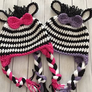 May include: Two crocheted zebra hats with black and white stripes, pink and purple bows, and braided tails.