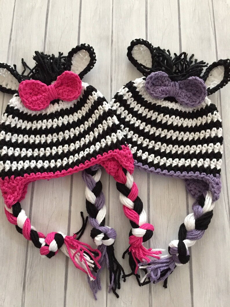 May include: Two crocheted zebra hats with black and white stripes, pink and purple bows, and braided tails.