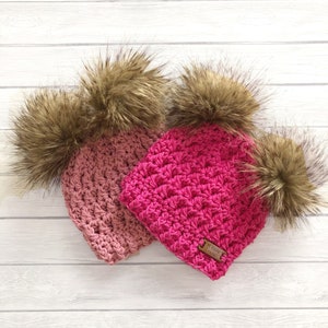 May include: Two crocheted hats with faux fur pom poms. One hat is pink and the other is a light pink color. Both hats have a brown leather tag with the text "My Roos Crochet" on them.