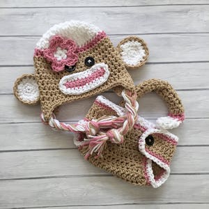 May include: A crocheted monkey hat and diaper set for a baby. The hat is brown, white, and pink with a flower on top. The diaper is brown, white, and pink with a pink trim.