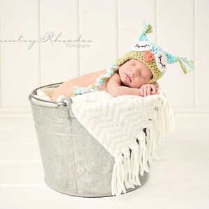 May include: A newborn baby wearing a crocheted owl hat sleeps in a metal bucket lined with a white blanket. The baby is wearing a green and blue owl hat with a white blanket.