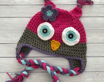 May include: A crocheted owl hat with a pink and gray body, white eyes with blue accents, a yellow beak, and a purple flower. The hat has a long braided cord with pink, purple, and blue strands.