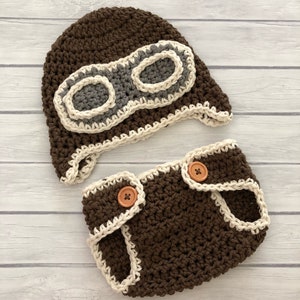 May include: A brown crochet baby pilot hat and diaper cover set. The hat features gray and off-white goggles. The diaper cover has two wooden buttons and off-white trim. The set is handmade.