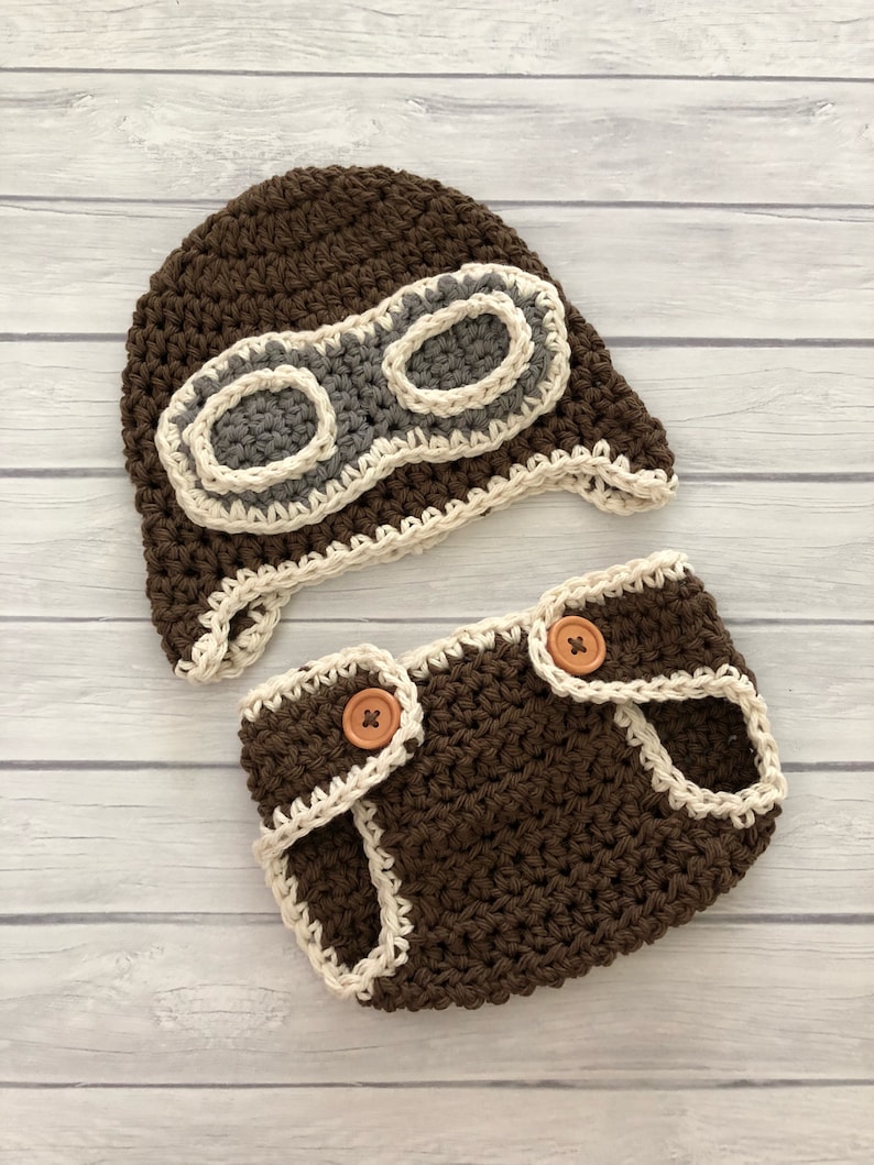 May include: A brown crochet baby pilot hat and diaper cover set. The hat features gray and off-white goggles. The diaper cover has two wooden buttons and off-white trim. The set is handmade.