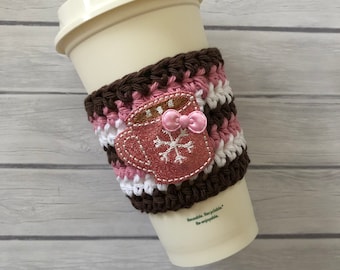 Op de afbeelding: Een witte koffiebeker met een gehaakte hoes in bruin, roze en wit. De hoes heeft een roze en wit sneeuwvlokkenontwerp met een bruine omtrek en twee roze strikken.