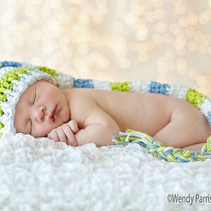May include: A newborn baby wearing a green, white, and blue crocheted hat and matching scarf sleeps on a white fluffy blanket. The background is a blurry bokeh of light.