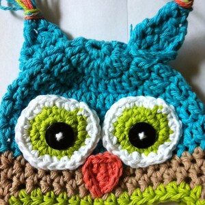 May include: A crocheted owl hat with green, white, brown, and blue yarn. The hat has two large eyes with black buttons and a small heart-shaped nose.