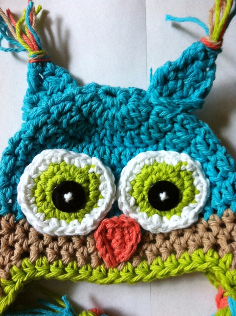 May include: A crocheted owl hat with green, white, brown, and blue yarn. The hat has two large eyes with black buttons and a small heart-shaped nose.
