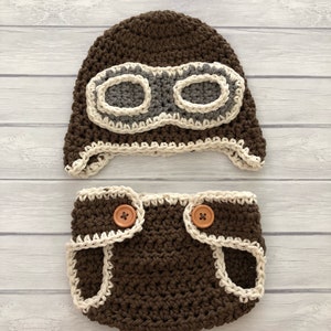 May include: A brown crochet baby pilot hat and diaper cover set. The hat features gray goggles with a cream-colored border. The diaper cover has cream-colored trim and wooden buttons. The set is designed for a newborn.