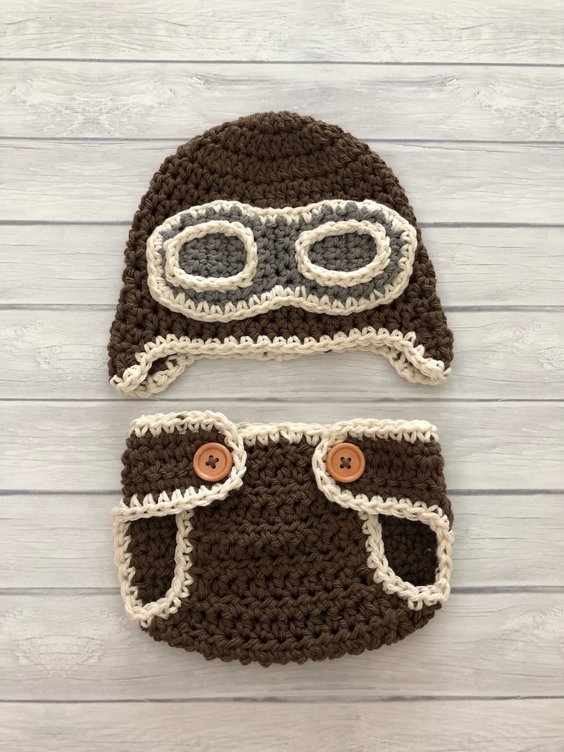 May include: A brown crochet baby pilot hat and diaper cover set. The hat features gray goggles with a cream-colored border. The diaper cover has cream-colored trim and wooden buttons. The set is designed for a newborn.