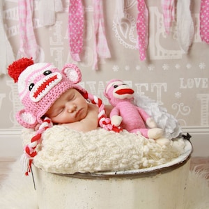 May include: A newborn baby wearing a pink and white crocheted monkey hat with a red pom-pom sleeps in a white wicker basket with a pink crocheted monkey toy.