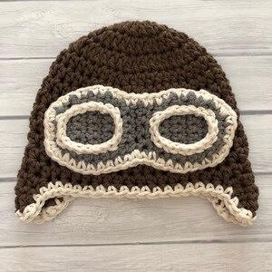 May include: A handmade brown crochet aviator hat with gray goggles and cream-colored trim. The hat features ear flaps and a textured stitch pattern, perfect for a baby or toddler. The goggles are a prominent design element.