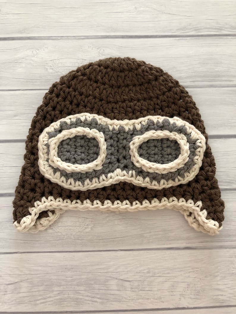 May include: A handmade brown crochet aviator hat with gray goggles and cream-colored trim. The hat features ear flaps and a textured stitch pattern, perfect for a baby or toddler. The goggles are a prominent design element.