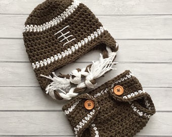 Puede incluir: Un gorro de fútbol de crochet marrón con costuras blancas y un cubrepañal de crochet marrón a juego con ribetes blancos y botones marrones.