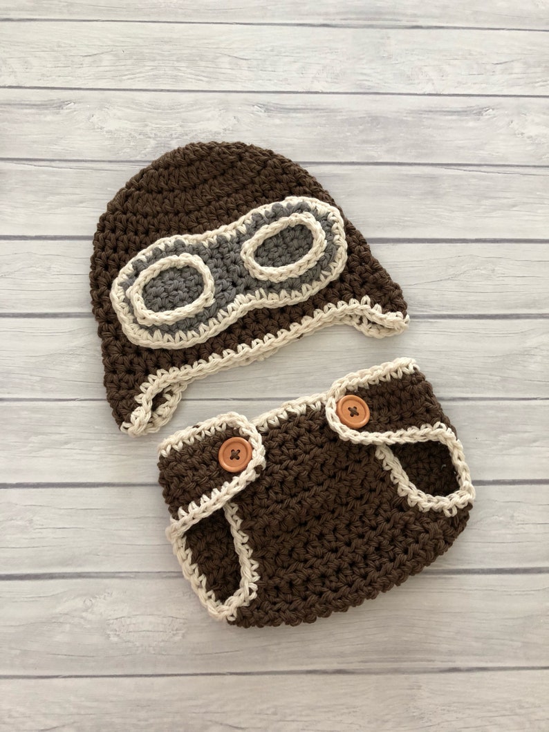 May include: A handmade crochet baby set in brown, featuring a pilot hat with goggles and matching diaper cover. The hat has a cream and gray goggle design. The diaper cover has wooden buttons and cream trim.