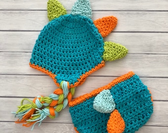 May include: A teal blue crocheted dinosaur hat and nappy set with orange, green and light blue accents. The hat has a long braided tail.