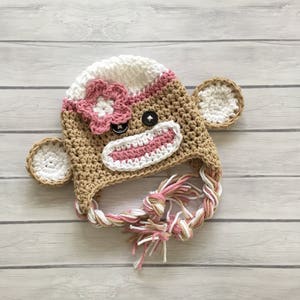 May include: A crocheted monkey hat with pink and white stripes, a pink flower, and brown yarn. The hat has a long, braided tie.