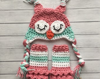 May include: A pink and green crocheted owl hat and matching fingerless mittens. The hat has a white face with black eyes and a small orange beak. The mittens have a pink and green striped pattern.