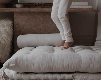 May include: A child jumps on a light gray, tufted floor cushion with a matching bolster pillow. The cushion is layered on top of a second, similar cushion. The cushion is on a light gray rug.
