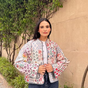 Puede incluir: Chaqueta colorida con estampado floral, base blanca y ribetes negros. La chaqueta presenta varios diseños florales en rojo, morado y naranja. La modelo lleva vaqueros azul oscuro.