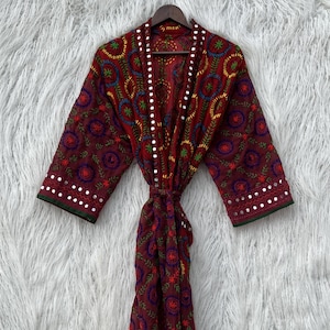 Può includere: Un accappatoio bordeaux ricamato con motivi floreali colorati e piccoli dettagli a specchio. L'accappatoio ha maniche lunghe e chiusura con cintura. Il capo è esposto su una gruccia di legno su uno sfondo bianco e soffice.
