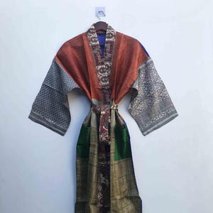 May include: A long, patterned robe with a belt. The robe features a mix of colors, including rust, gray, green, and gold. The sleeves and belt have intricate designs. The robe is hanging on a wooden hanger against a white wall.