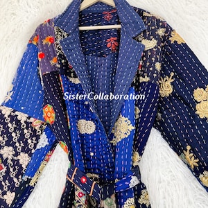 May include: A patchwork robe in shades of blue, with embroidered floral and geometric patterns. The robe features a shawl collar, a matching belt, and is hung on a wooden hanger. The text "SisterCollaboration" is visible on the robe.